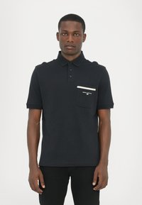 Svart polo skjorte med krage, tre knapper foran og lomme med kontrastfarget hvit stripe. Mykt bomullsstoff med klassisk passform.