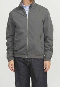 Gray zip-up jacket with a stand collar, side pockets, and ribbed cuffs. Smooth fabric and a fitted design, paired with dark jeans.