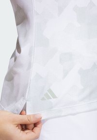 Hvid atletisk top med et subtilt gråt camouflage-mønster. Tekstureret stof med et logo og tre grønne striber i kanten.