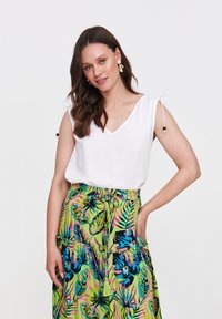 Biała bluzka bez rękawów z dekoltem w kształcie litery V i detalami ze sznurkiem; zestawiona z zieloną spódnicą z intensywnymi tropikalnymi wzorami liści i ozdobnymi frędzlami.