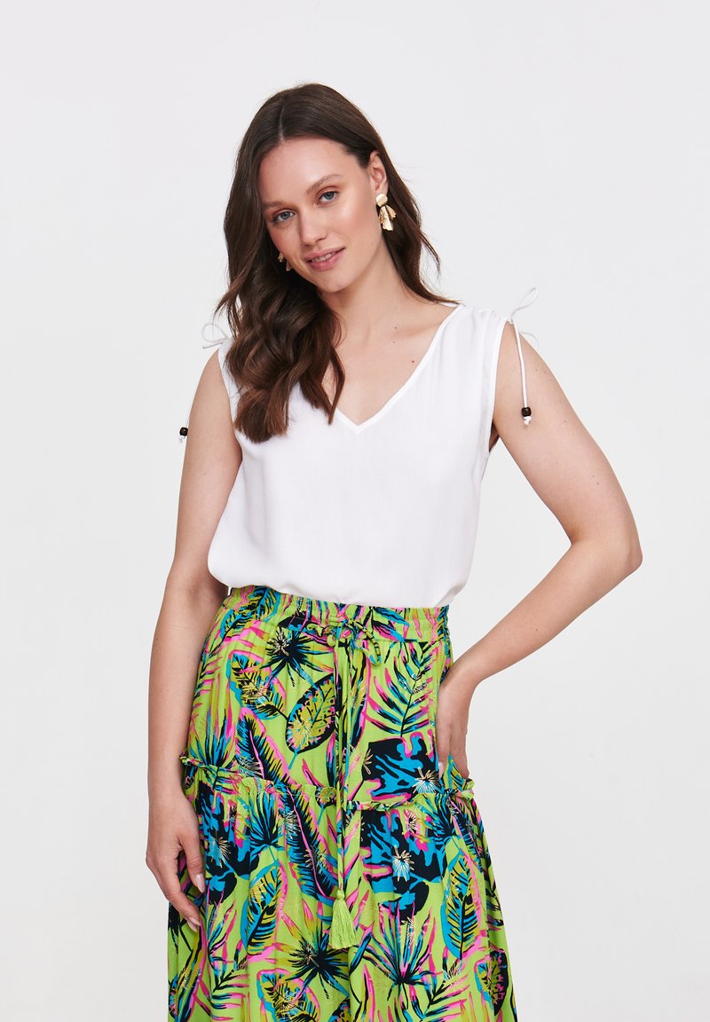 Biała bluzka bez rękawów z dekoltem w kształcie litery V i detalami ze sznurkiem; zestawiona z zieloną spódnicą z intensywnymi tropikalnymi wzorami liści i ozdobnymi frędzlami.