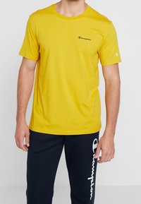 Žluté bavlněné tričko s kulatým výstřihem a krátkými rukávy; na hrudi se nachází malé černé vyšívané logo Champion.