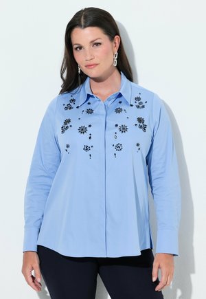 Kvinde med mørkt hår bærer en lyseblå bluse med lange ærmer dekoreret med sorte blomsterprint og mørke bukser mod en ensfarvet baggrund.
