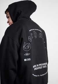 Black hoodie with a large hood, featuring white text and graphic prints on the back. Soft fabric with a relaxed fit.