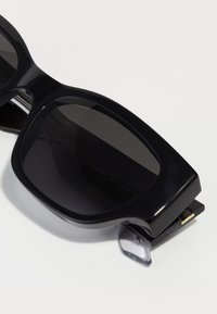 Store solbriller i sort med mørke linser, tykke stel og en blank finish. Bemærkelsesværdig gylden detalje ved tempelpunktet.