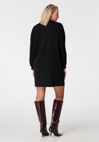 Robe noire à manches longues avec une coupe droite, associée à des bottes en cuir bordeaux gaufré crocodile montant jusqu'aux genoux, avec une finition mate douce.