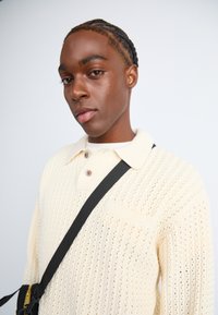 Jersey de polo de color crema, tejido, con un patrón texturizado, un collar y un único botón. Combinado con una correa de bolsa cruzada negra.