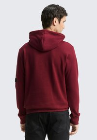 Felpa maroon con tasca a marsupio, cappuccio regolabile con lacci e polsini a coste. Vista posteriore che mostra texture e vestibilità. Jeans neri sotto.