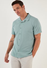 Mintgroene polo met korte mouwen, gemaakt van een gestructureerde stof, met een knoopsluiting aan de voorkant en een gespreide kraag. De polo heeft een rechte zoom en versterkte naden.