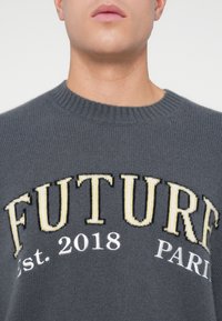 Uomo che indossa un maglione lavorato a maglia grigio con grande testo giallo e bianco che scrive "FUTURE Est. 2018 PARIGI" sul petto.