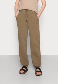 Pantalones de chándal marrones con cintura ajustable, que cuentan con bolsillos laterales y puños elásticos. Combinados con sandalias negras sobre un fondo neutro.