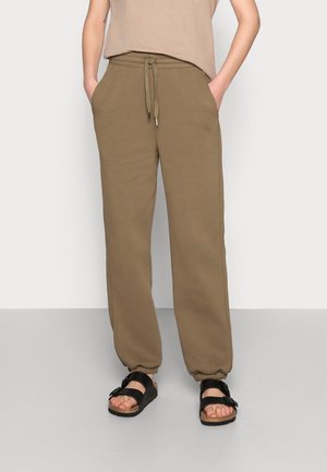 Pantalons de survêtement marron avec taille à cordon de serrage, comportant des poches latérales et des poignets élastiques. Associés à des sandales noires sur un fond neutre.
