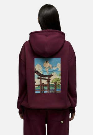 Personne aux longs cheveux foncés porte un sweat à capuche bordeaux représentant un lutteur de sumo faisant du skateboard au-dessus d'une porte torii japonaise traditionnelle sous un ciel bleu.