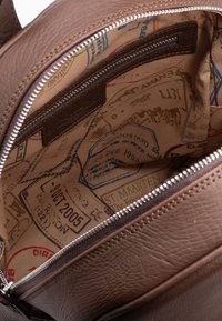 Interno di una borsa in pelle marrone con una texture liscia, dotato di una tasca con zip e una fodera in tessuto decorata con timbri di viaggio e date.