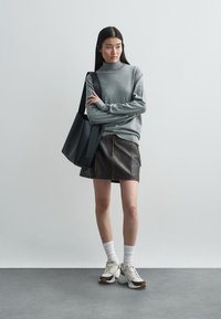 Szary golf, brązowa skórzana spódnica z widocznym przeszyciem, białe skarpetki i masywne sneakersy. Nosi czarną torbę na ramię.