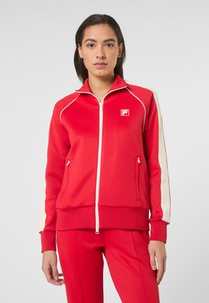 Ung kvinde iført en rød lynlåsjakke med hvide striber på ærmerne og matchende røde bukser, stående foran en ensfarvet baggrund.