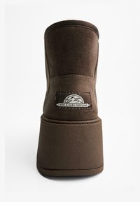 Stivaletto marrone con tomaia simile alla pelle scamosciata e suola piatta spessa e texturizzata. Caratterizzato da una patch con logo vicino al tallone e dettagli cuciti.