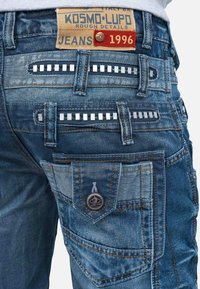 Jeans in denim blu con una texture ruvida, dotati di borchie metalliche sulle tasche posteriori, cuciture arancioni e un'etichetta con la scritta "KOSMO•LUPO 1996."