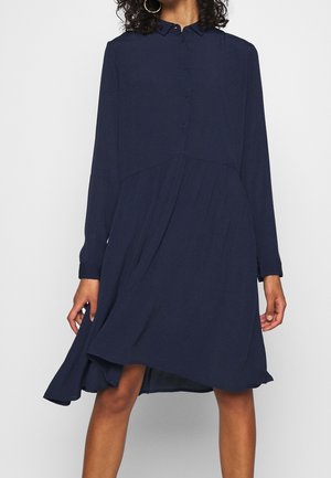 Kobieta ubrana w granatową sukienkę na guziki z długim rękawem, o asymetrycznym dole oraz kolczykach kołach, stojąca na jednolitym tle.