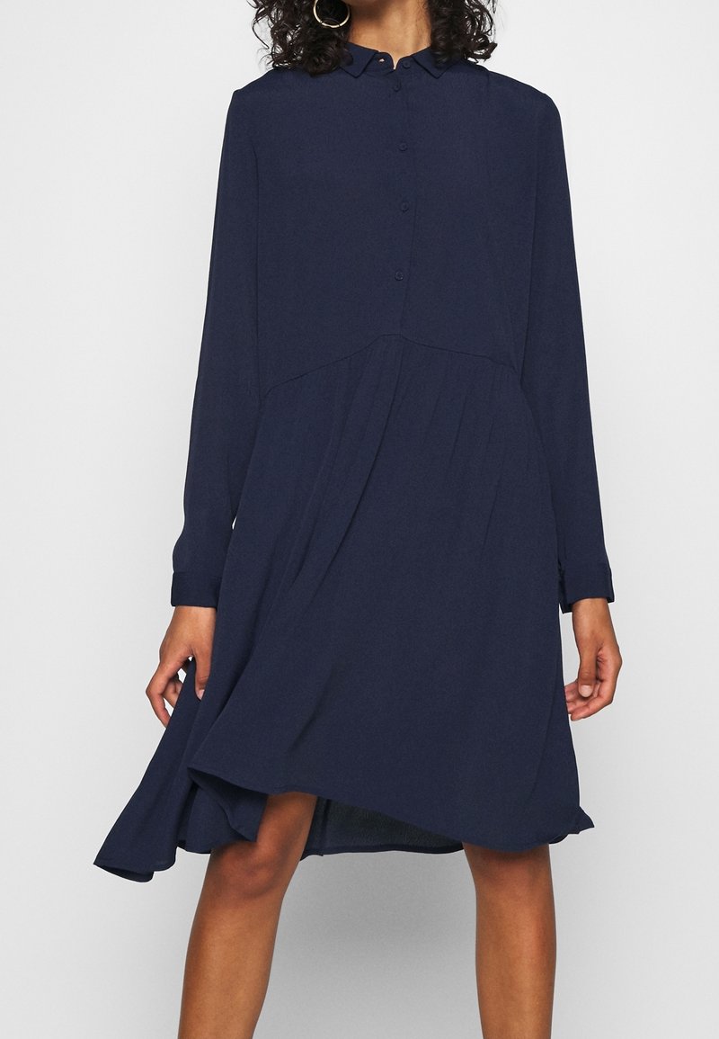 Femme portant une robe chemise bleu marine à manches longues avec un ourlet asymétrique et des boucles d'oreilles créoles, debout devant un fond uni.