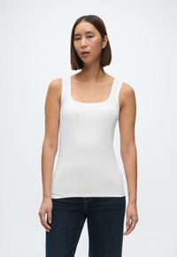 Biały dopasowany top na ramiączkach z kwadratowym dekoltem i szerokimi ramiączkami, wykonany z gładkiego materiału. Noszony z ciemnoniebieskimi dżinsami.
