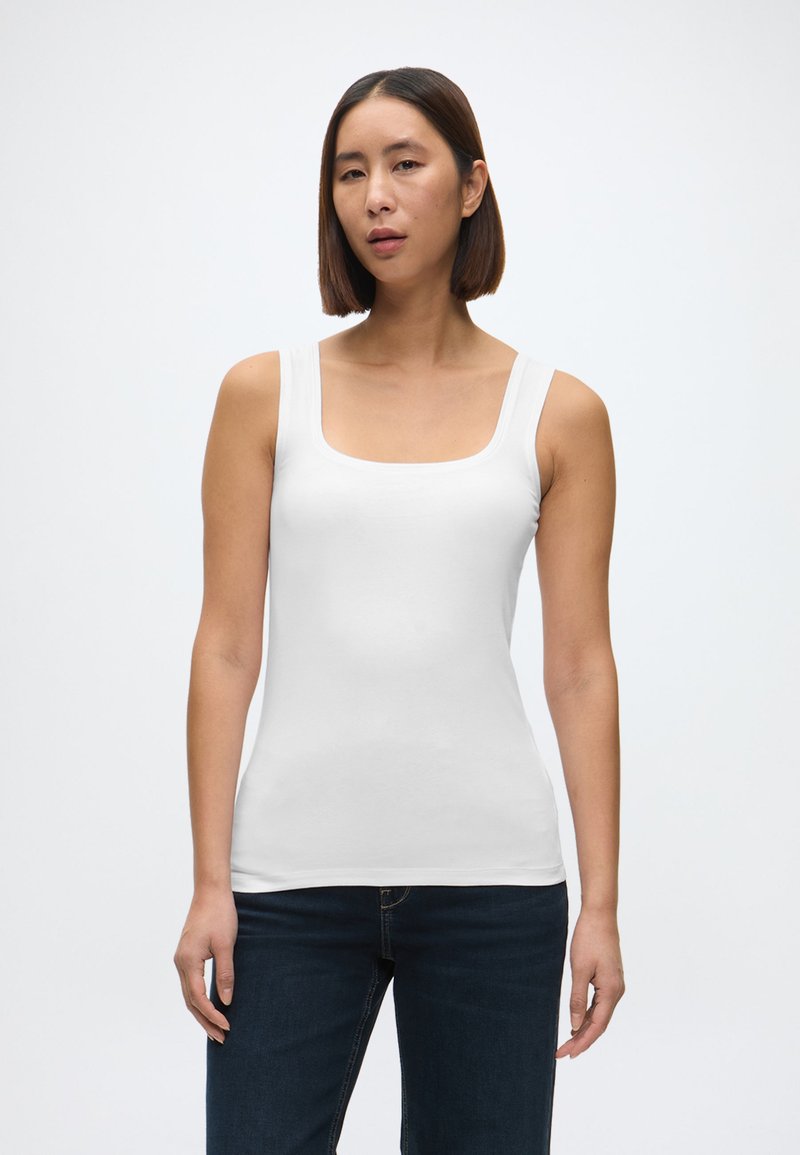 Biały dopasowany top na ramiączkach z kwadratowym dekoltem i szerokimi ramiączkami, wykonany z gładkiego materiału. Noszony z ciemnoniebieskimi dżinsami.