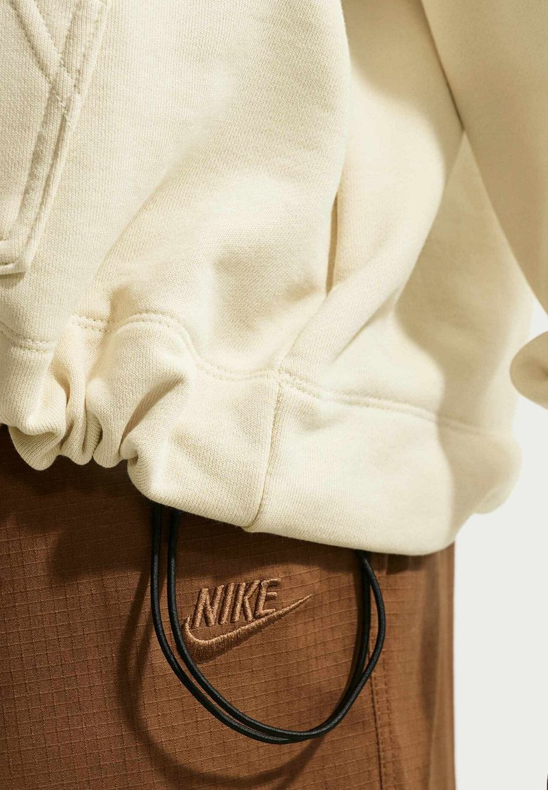 Personne portant un hoodie crème et un pantalon Nike marron avec un cordon noir pendant, seules les zones du torse et des hanches sont visibles.