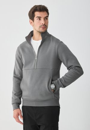 Šedá mikina na zips s vysokým golierom, so spodným švom a bočnými vreckami. Vyrobená z jemnej, hladkej látky.