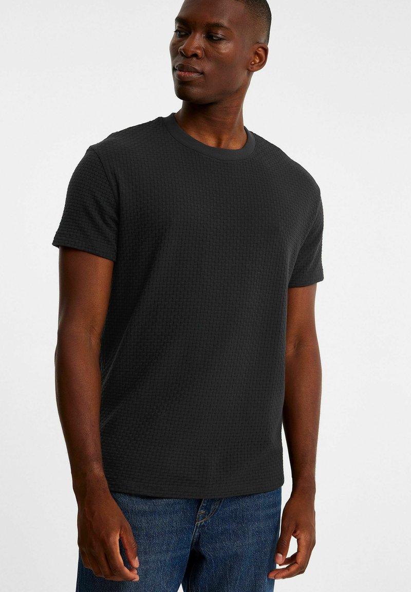 Sort tekstureret kortærmet T-shirt med rund halsudskæring, der har et subtilt geometrisk mønster og en afslappet pasform, parret med blå denimjeans.