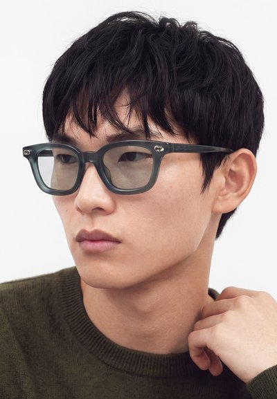 Jeune homme aux cheveux noirs courts portant des lunettes rectangulaires foncées et un pull vert olive, regardant légèrement sur le côté avec une expression neutre.