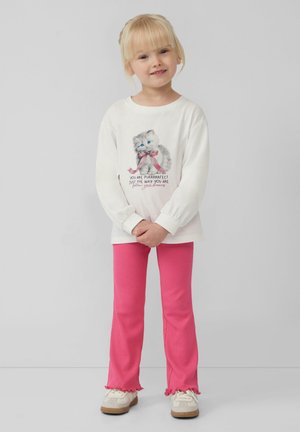 Camicia bianca a maniche lunghe con grafica di un gatto e testo, abbinata a pantaloni a campana rosa a coste con orli arricciati. Sneakers bianche completano l'outfit.