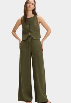 Vrouw draagt olijfgroene mouwloze top met knopen en bijpassende wijdvallende broek met hoge taille en trekkoord, staand met handen op heupen.