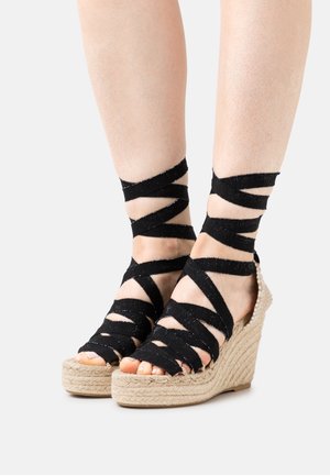 Sandales compensées noires en espadrille avec des sangles élastiques croisées, talon enveloppé de jute et design ouvert. Matériau texturé et accents décoratifs.