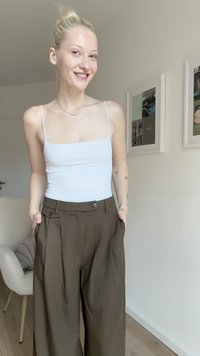 Witte, aansluitende tanktop met dunne bandjes gecombineerd met hoge, bruine, geplooide broeken. Glad textuur van de stof, twee knopen zichtbaar.