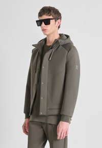 Olivgrüne Zip-Jacke mit Kapuze und Druckknöpfen; kombiniert mit einem passenden Rundhalsausschnitt-Shirt. Das Model trägt schwarze rechteckige Sonnenbrillen.