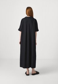 Vestido maxi de linho preto com mangas curtas com punho e corte reto, apresentando um yoke nas costas e bolsos laterais, usado com sapatos pretos tipo slip-on.