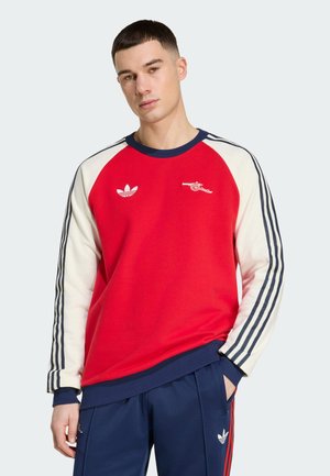 Sweat-shirt rouge avec manches crème et accents bleus, présentant un col rond, des logos brodés et des détails rayés sur les bras.