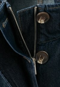 Jean en denim avec une fermeture éclair en métal argenté partiellement ouverte et deux grands boutons foncés à quatre trous sur la ceinture.