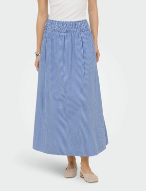 Femme portant une longue jupe vichy bleue et blanche avec une ceinture froncée et des chaussures plates tissées beige, debout devant un fond uni.