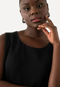 Schwarzes ärmelloses Oberteil mit rundem Ausschnitt und dezenten Falten. Das Modell trägt große silberne Creolen und einen neutralen Lippenstift.