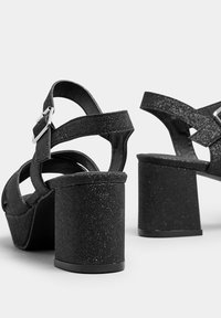 Sandalias negras de tacón cuadrado con textura brillante, que presentan correas entrelazadas y una hebilla de metal. Diseño de tacón robusto para mayor estabilidad.