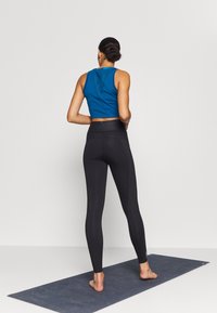 Modrý bezrukávový crop top spárovaný s černými legínami s vysokým pasem, oba fit, na šedé yoga podložce. Detaily textury viditelné na topu.