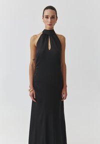 Musta halterneck-mekko, jossa on avonainen kaula-aukko. Sileä rakenne, vartalonmyötäinen siluetti ja lattiaan ulottuva muotoilu. Minimalistinen tyyli ilman kuvioita.