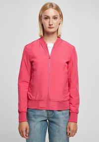 Roosa bomber-jakk, millel on tõmblukk ees, elastsed mansetid ja alumine äär, kerge materjal ja sile pind. Kombineeritud siniste teksadega.