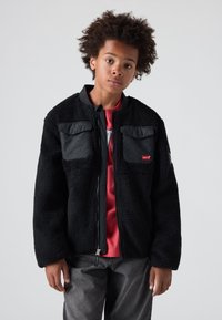 Crna flis jakna s dva siva džepa na prsima, zatvaranje na zip, i rebrasti okovratnik. Nosena preko crne majice kratkih rukava, u kombinaciji sa sivim hlačama.