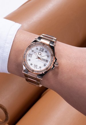 Reloj metálico con una correa de oro rosa, esfera blanca texturizada, números romanos y acentos similares a diamantes en el bisel.