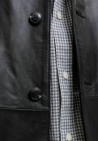 Gros plan d'une veste en cuir noire avec de grands boutons sur une chemise à carreaux blanche et noire boutonnée.