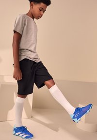 Blå fotbollsskor med ljusare blå detaljer, grå T-shirt, svarta shorts och vita knälånga strumpor, stående i en neutral miljö.