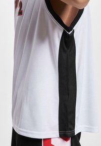 Maillot de basket blanc avec une bande verticale noire et un numéro rouge partiellement visible sur le côté, porté par une personne sur un fond uni.
