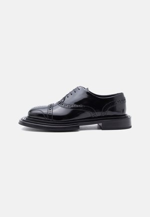 Zapato de vestir brogue de cuero negro pulido con perforaciones en forma de ala y tacón apilado, mostrado de perfil sobre un fondo blanco.